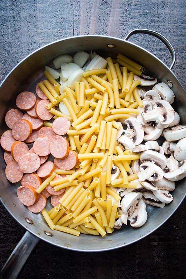 Skillet with dry ziti pasta, sliced sausages, sliced mushrooms, and onions.