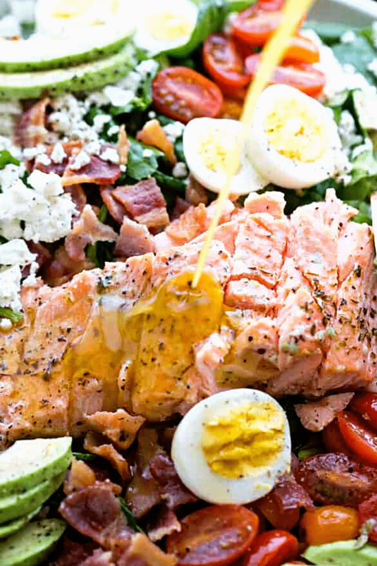 pouring salad dressing over a salmon fillet surrounded by halved boiled eggs, crumbles of bacon, salad greens, cherry tomatoes, and sliced avocado.