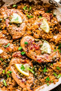 A top view of Chicken Piccata and Rice in a pan with capers, parsley, and sliced lemon