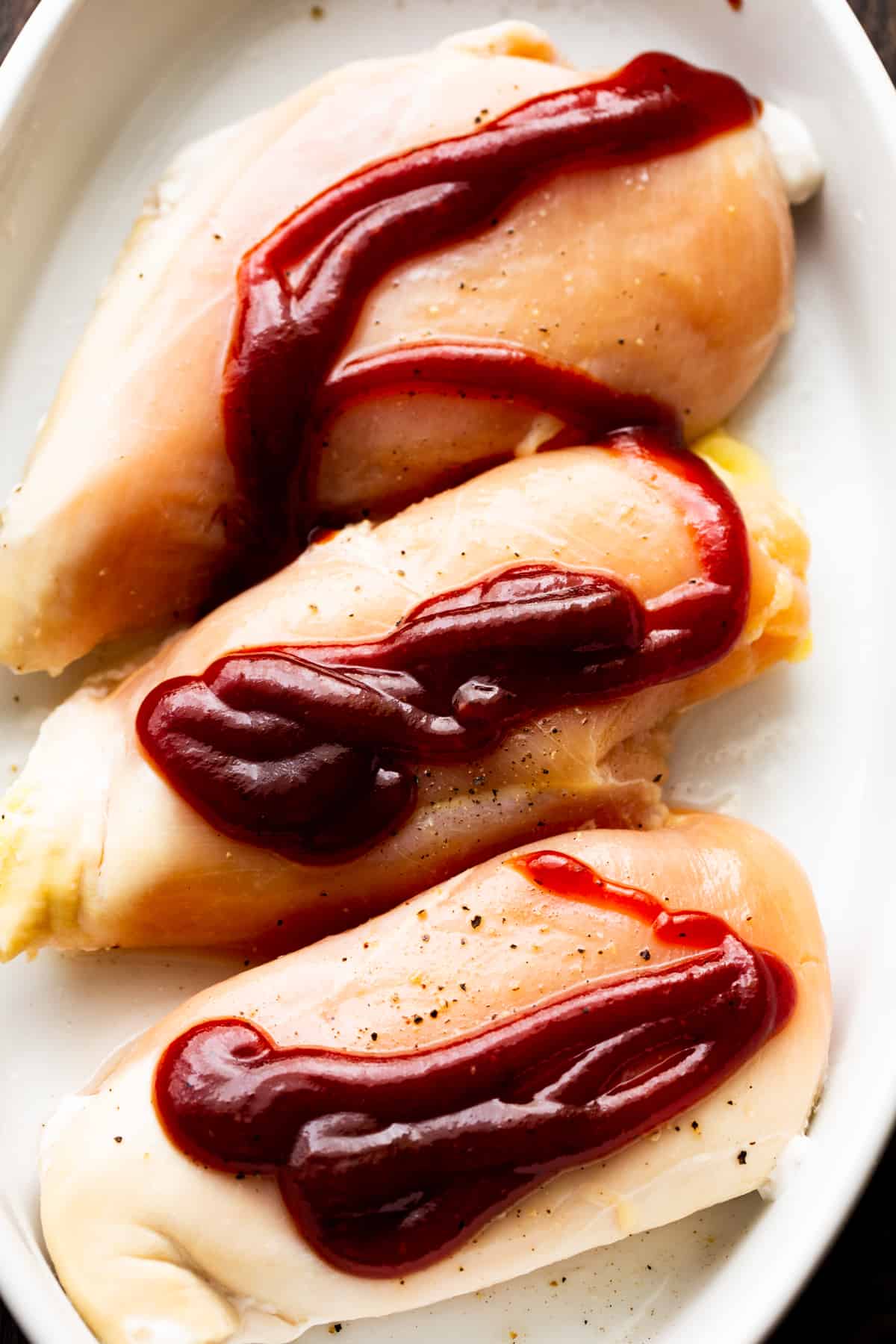 overhead photo of three raw chicken breasts in a baking dish, topped with barbecue sauce.