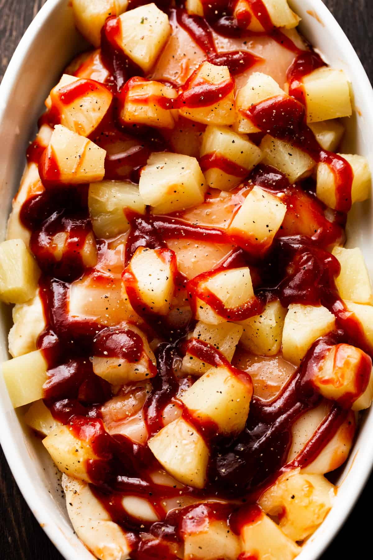 three raw chicken breasts in an oval dish, topped with pineapple chunks and bbq sauce.