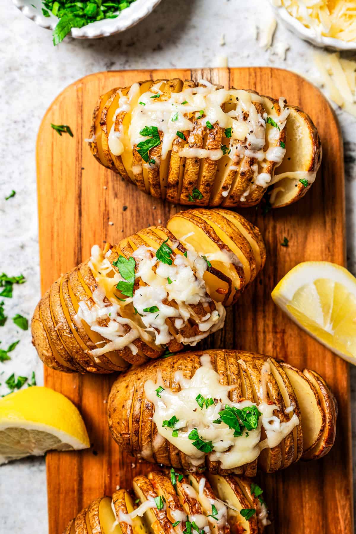 Overhead view of hasselback potatoes topped with melted parmesan on a wooden cutting board, next to lemon wedges.