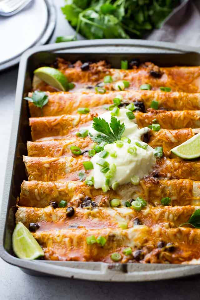 Ground Turkey Black Bean Enchiladas in a gray-colored baking pan and topped with green onions, sour cream, and lime wedges.