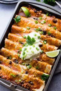 Ground Turkey Black Bean Enchiladas arranged in a gray-colored baking pan and topped with green onions, sour cream, and lime wedges.
