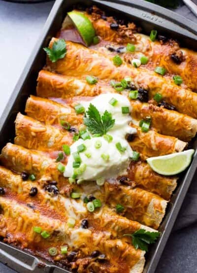 Ground Turkey Black Bean Enchiladas arranged in a gray-colored baking pan and topped with green onions, sour cream, and lime wedges.