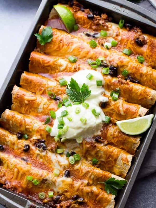 Ground Turkey Black Bean Enchiladas arranged in a gray-colored baking pan and topped with green onions, sour cream, and lime wedges.