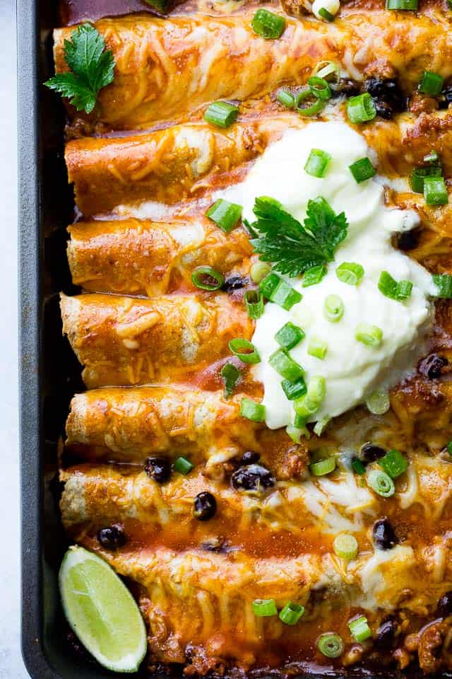 Ground Turkey Enchiladas arranged in a gray-colored baking pan and topped with green onions, sour cream, and lime wedges.