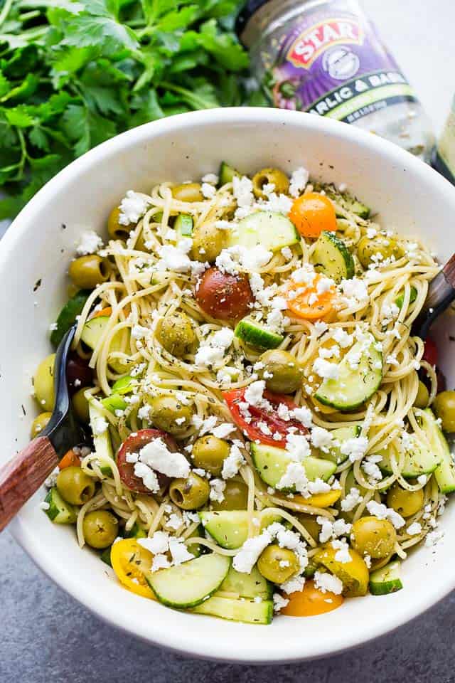 Overhead shot of a large salad bowl with spaghetti, diced cucumbers, chopped tomatoes, whole olives, and crumbled feta cheese.