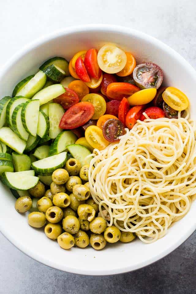 Overhead image of the ingredients needed for making a spaghetti salad. There's sliced cucumbers, halved grape tomatoes, and whole green marinated olives.