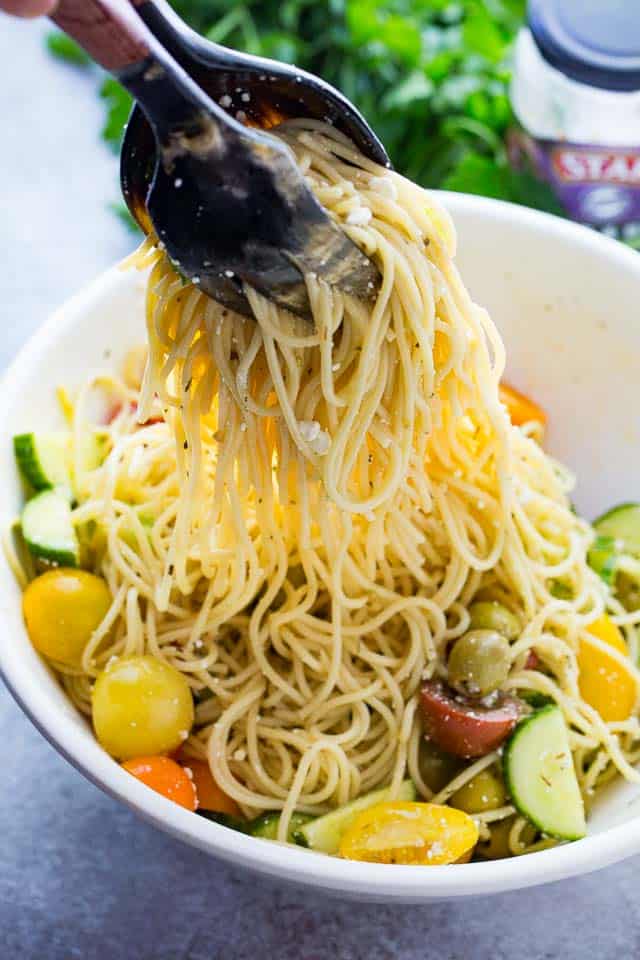 Two serving spoons tossing spaghetti with fresh veggies.