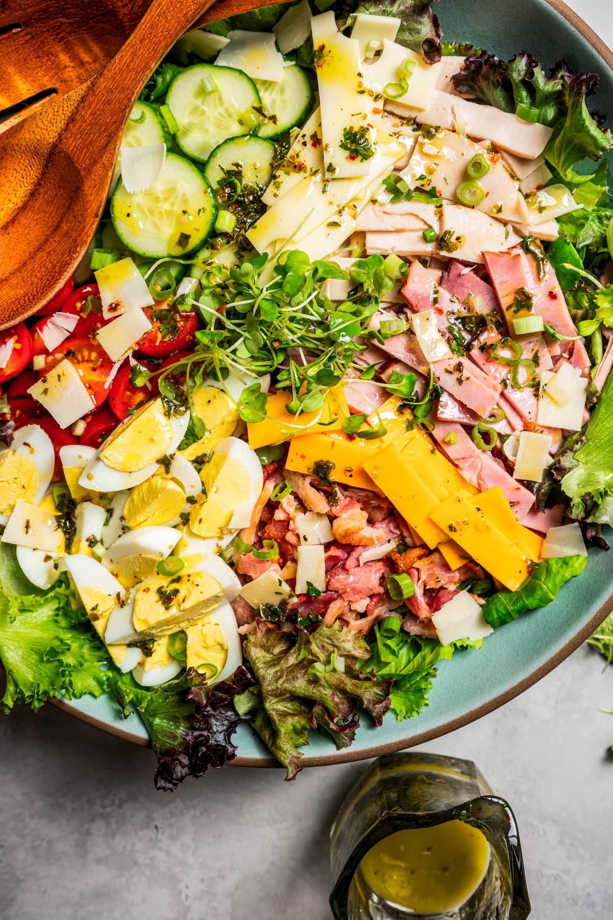 Chef salad in a large salad bowl dressed in Italian dressing next to wooden salad tongs.