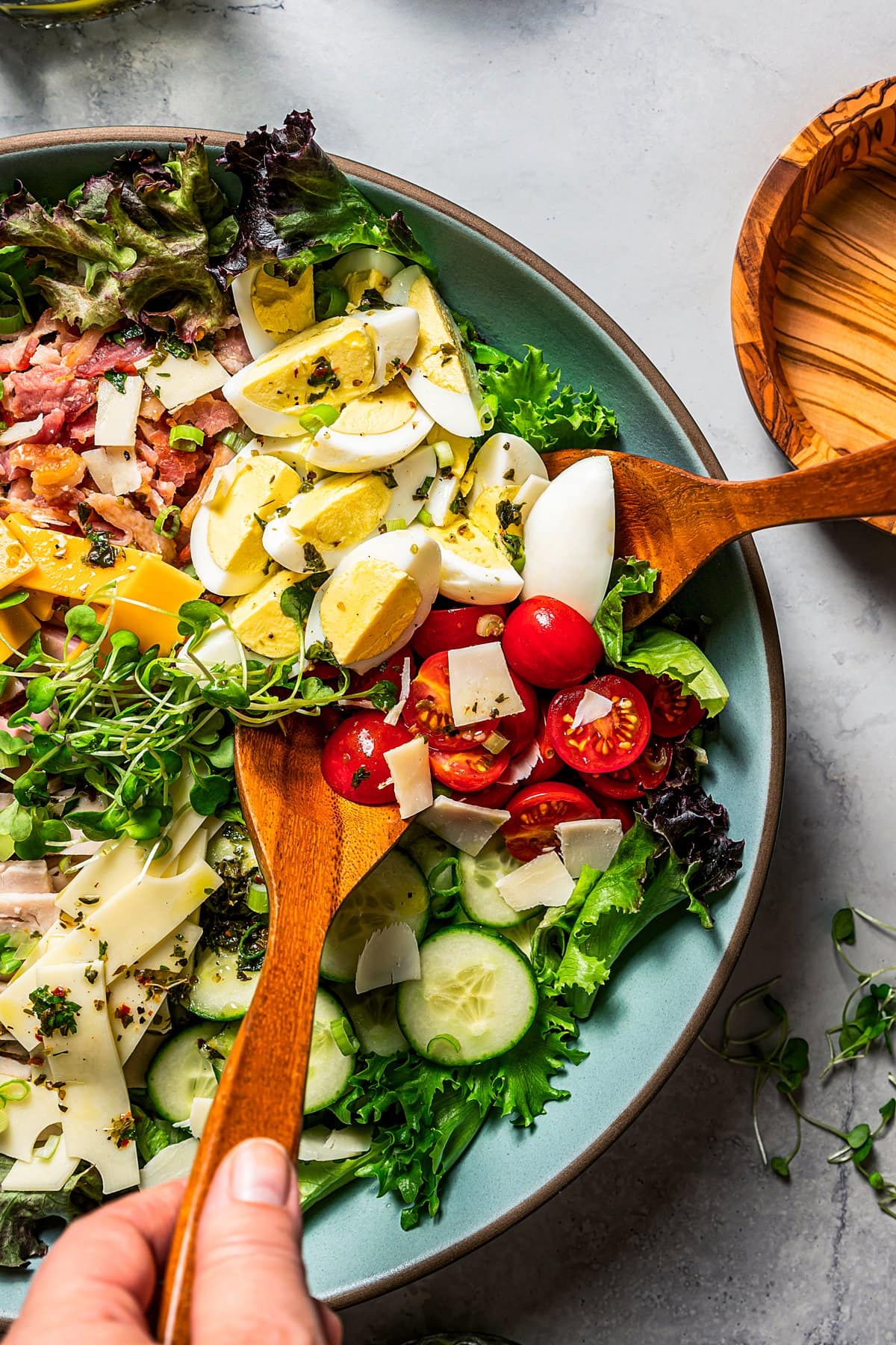 Hands use wooden tongs to toss a chef salad in a large bowl.