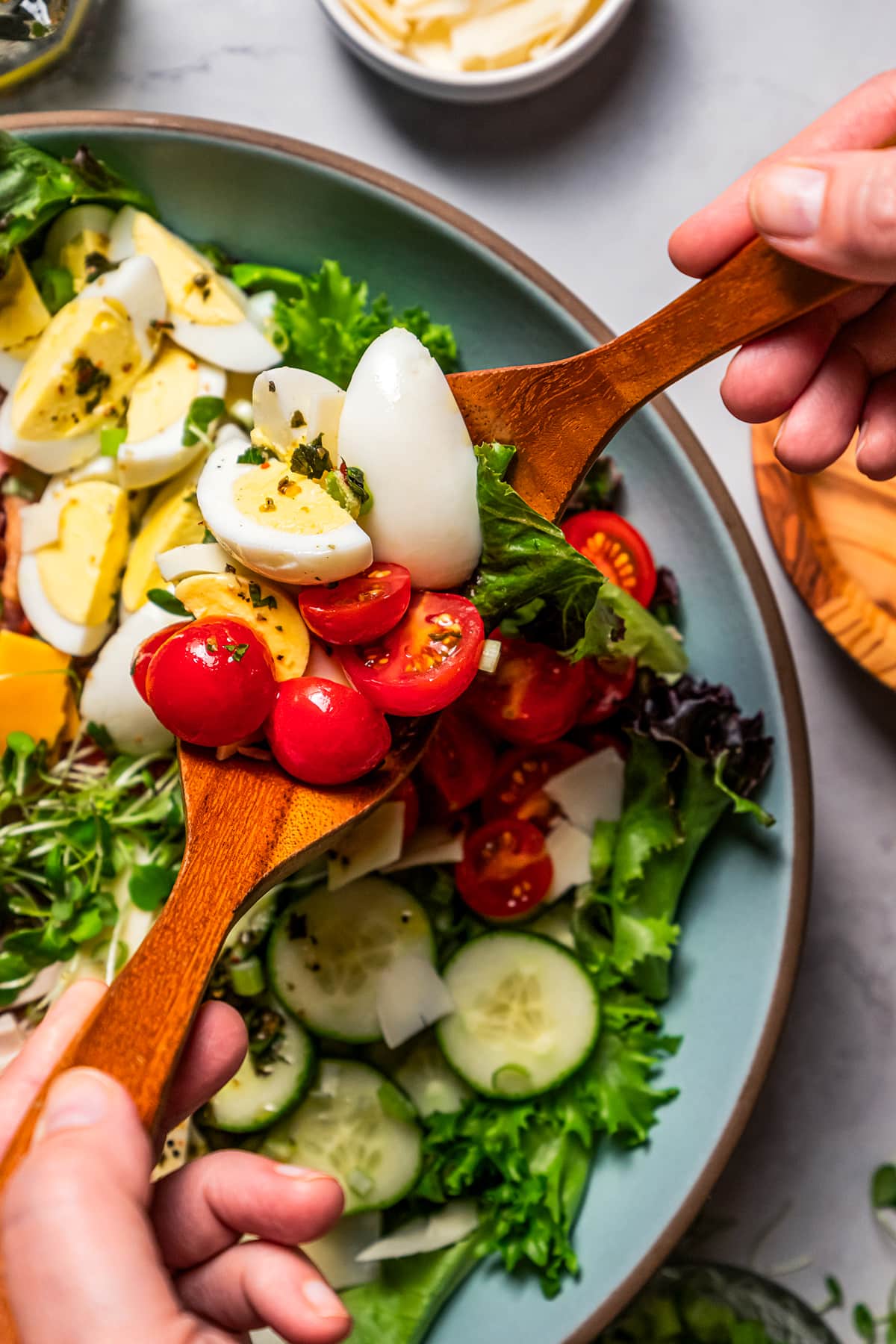 Close up of two hands using wooden salad tongs to toss a chef salad in a large bowl.