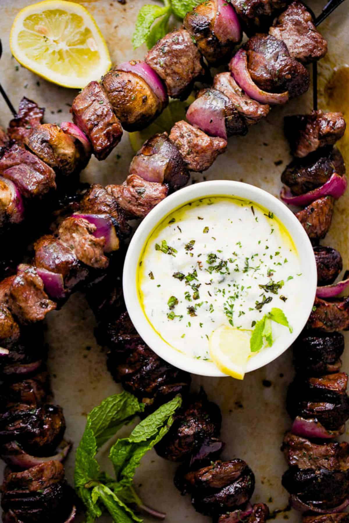 Steak kebabs on a baking sheet with a bowl of dip set in the middle of the skewers.