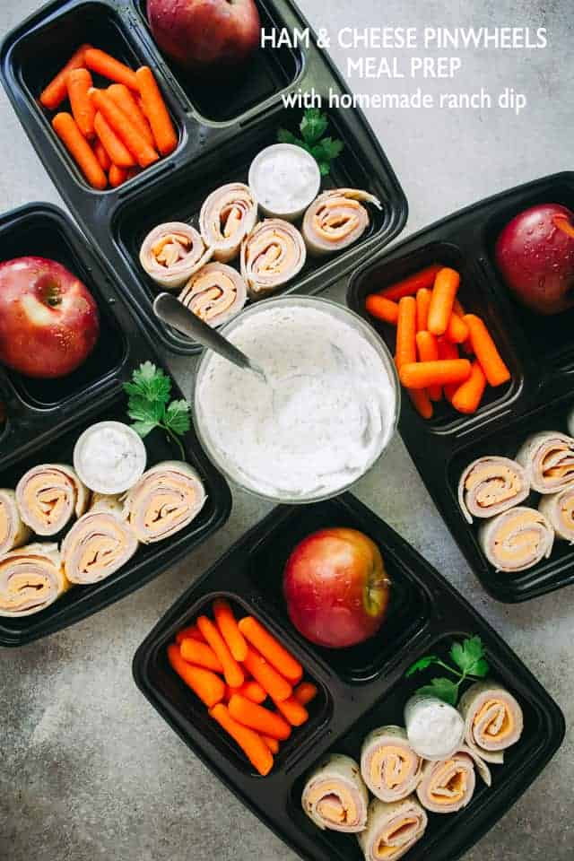 Black containers filled with ham and cheese pinwheels, baby carrots, and apples. There's a bowl of ranch dip in the middle of the containers.