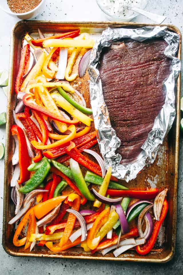 Seasoned flank steak wrapped in foil next to sliced veggies on a sheet pan.
