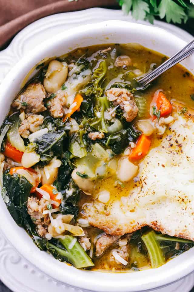 Italian Sausage Soup with kale and beans served in a bowl with a slice of crusty bread.