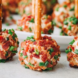 A close-up of mini cheeseballs sprinkled with bacon and chives and a pretzel stick going through the center of each cheeseball.