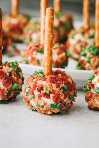 A close-up of mini cheeseballs sprinkled with bacon and chives and a pretzel stick going through the center of each cheeseball.