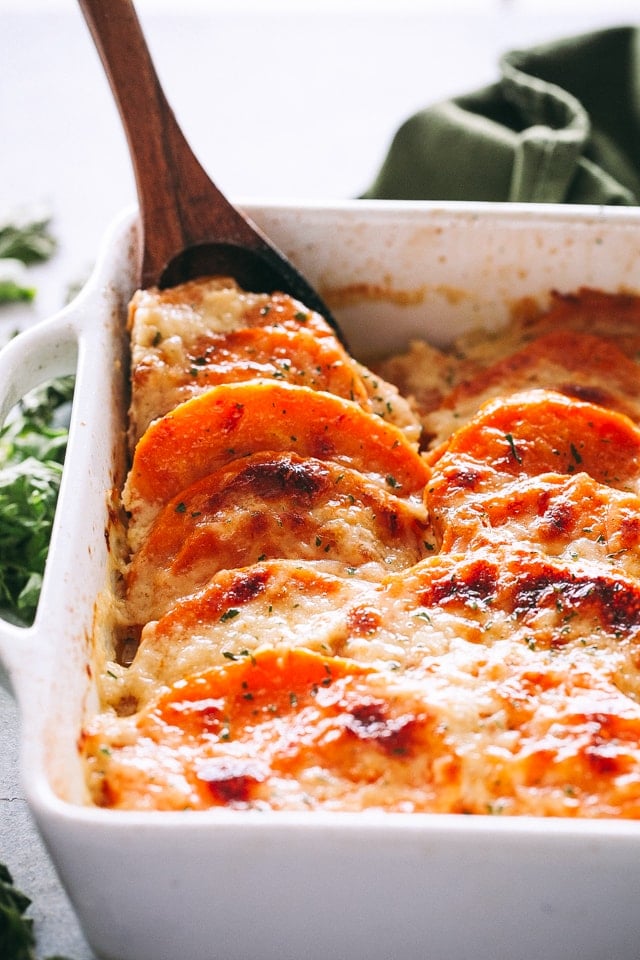 Wooden spoon lifting out a serving of sweet potato gratin from a baking dish.