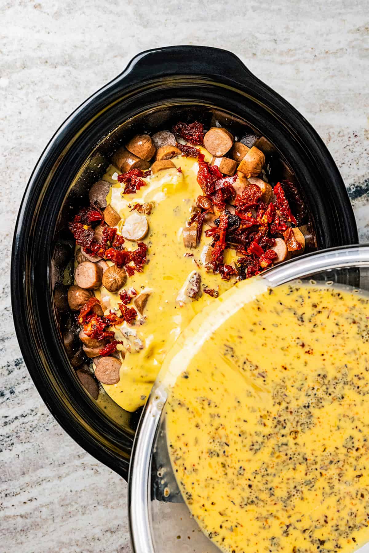 Pouring beaten eggs over breakfast sausages in a slow cooker insert.