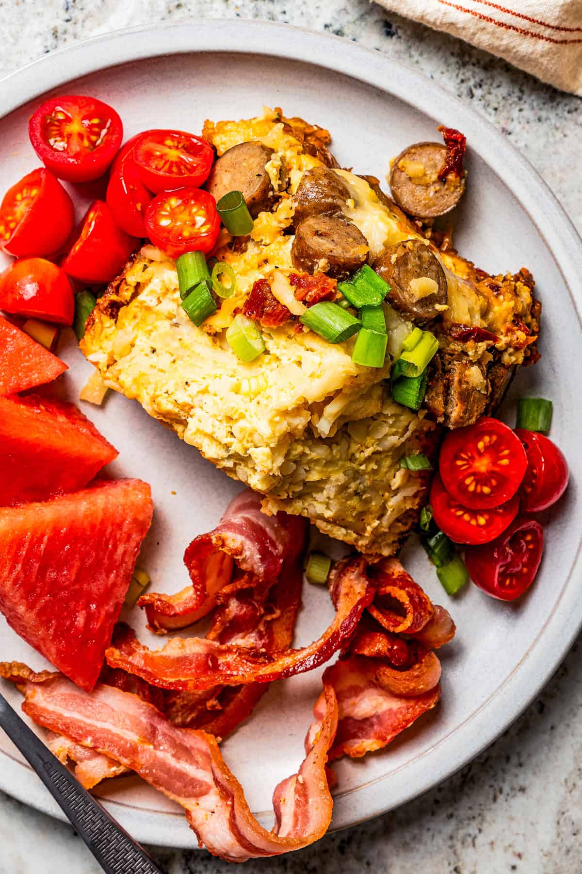 Slice of crockpot breakfast casserole served on a plate with cherry tomatoes, bacon, and watermelon triangles.