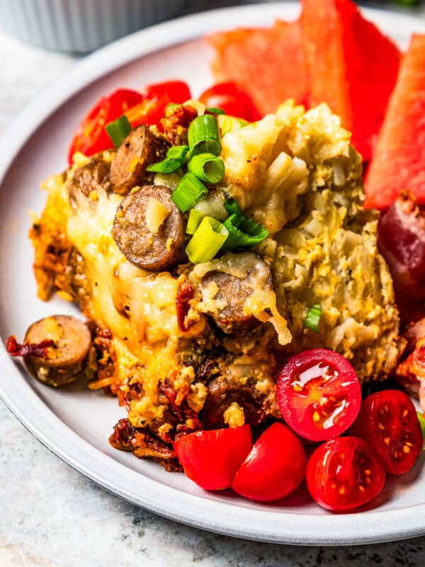 A serving of slow cooker breakfast casserole next to sliced cherry tomatoes, watermelon triangles, and crispy bacon on a white plate.