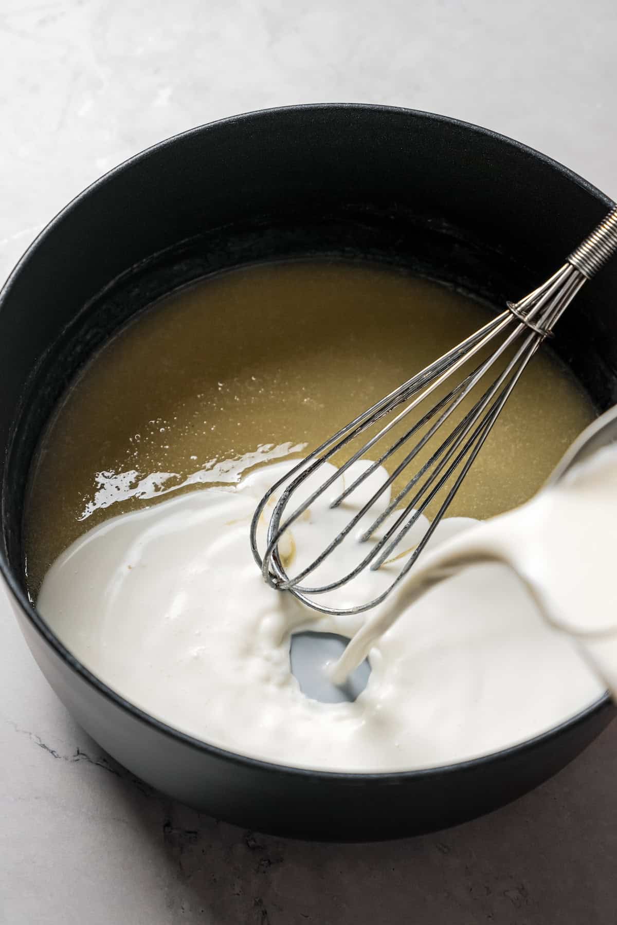 Milk being poured into a pot and whisked with melted butter and flour to make a roux.