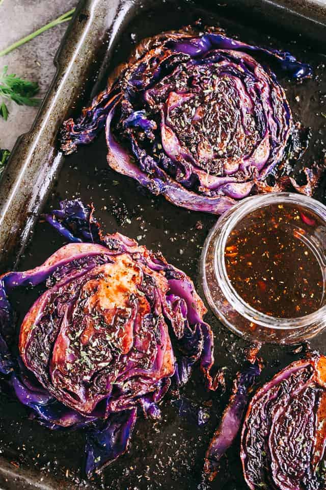 Three Roasted Cabbage Steaks arranged on a dark baking sheet, with a jar of balsamic glaze set in the center of the sheet.