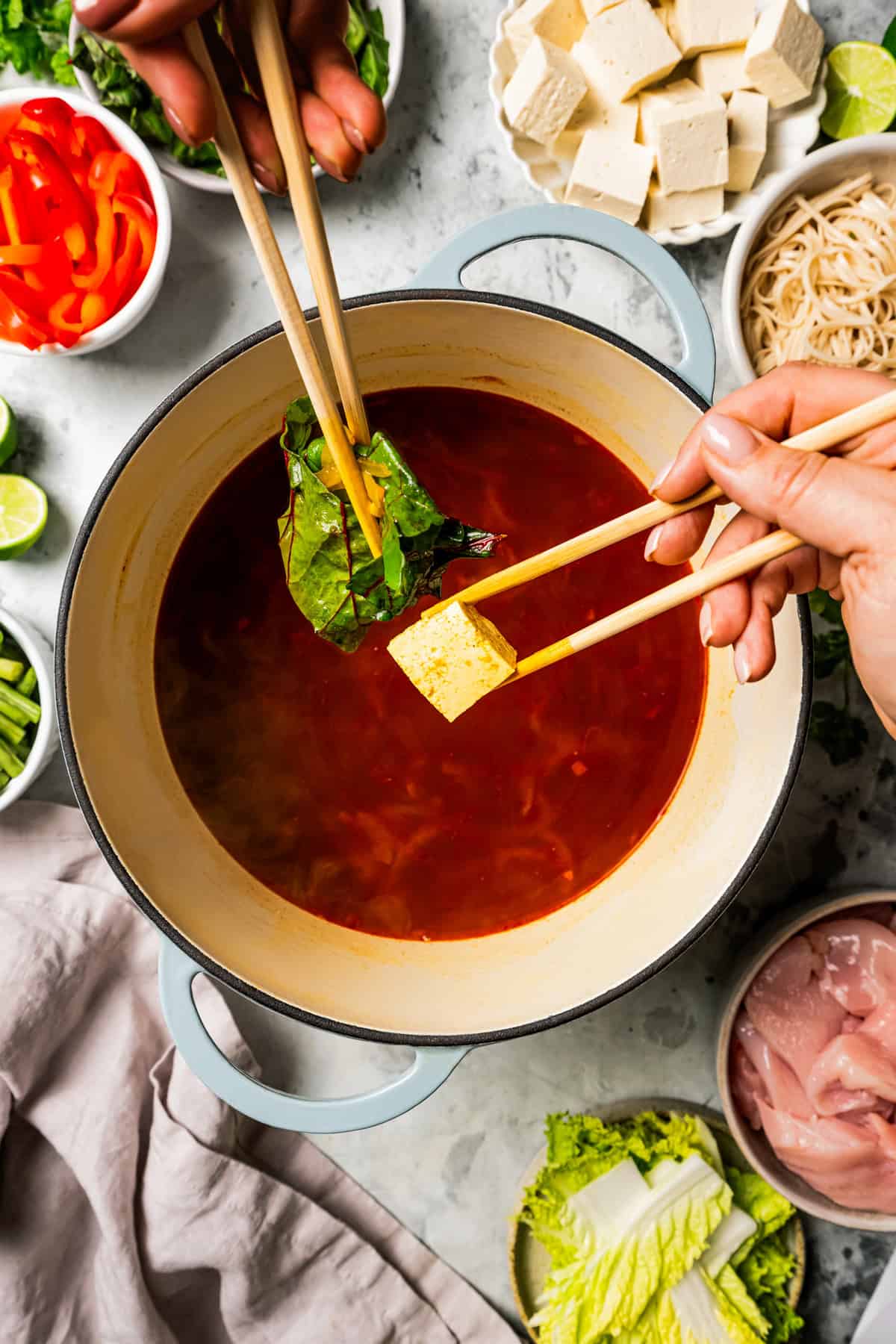 Overhead view of hands holding chopsticks with tofu and cabbage over a Chinese hot pot surrounded by bowls of ingredients.