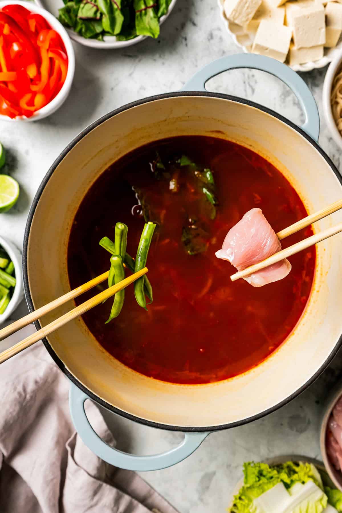 Overhead view of one set of chopsticks holding chicken and another set holding green beans over a Chinese hot pot surrounded by bowls of ingredients.