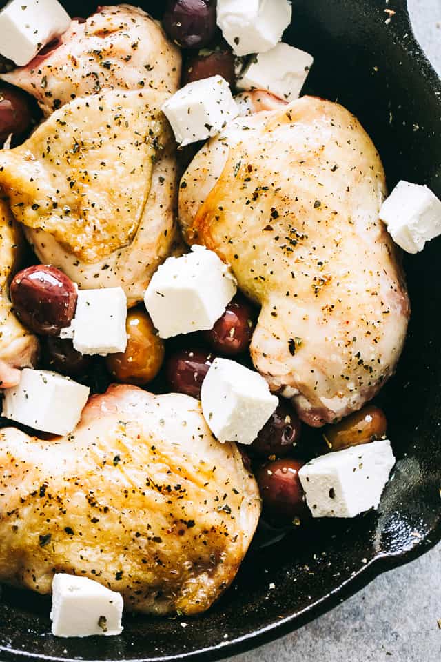 Browned chicken thighs in a skillet with cubes of feta cheese and olives.