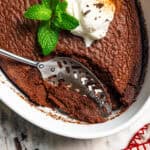 Chocolate fudge cake garnished with whipped cream and mint leaves, with a spoon in an oval baking dish.