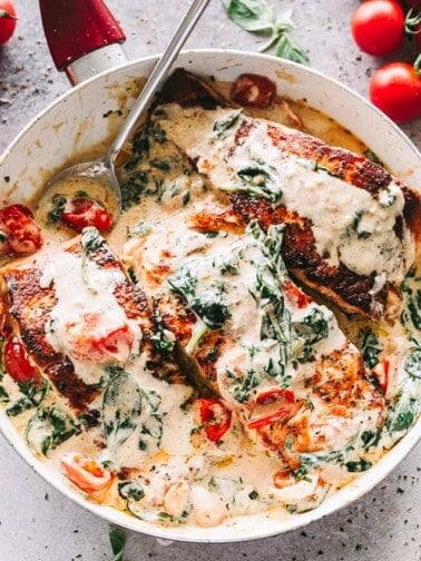 Overhead image of a skillet with three salmon fillets in a cream sauce mixed with baby spinach and cherry tomatoes.