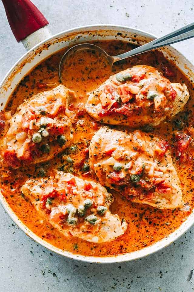 Zoomed out overhead image of chicken in tomato sauce in a skillet with a serving spoon.