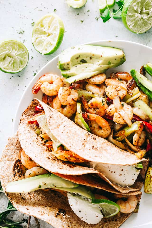 Overhead view of shrimp fajitas in tortillas, with sliced avocado and an extra serving of shrimp and vegetables on a white plate.