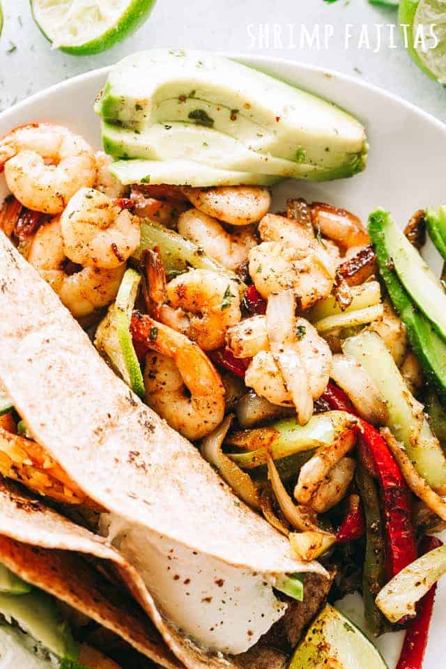 Overhead view of shrimp fajitas in tortillas, with sliced avocado and an extra serving of shrimp and vegetables on a white plate.