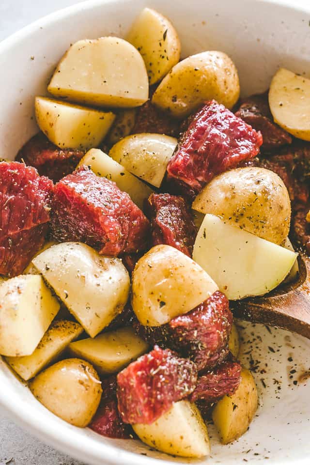 Uncooked steak bites and chopped potatoes marinating with garlic and herbs in a white bowl.