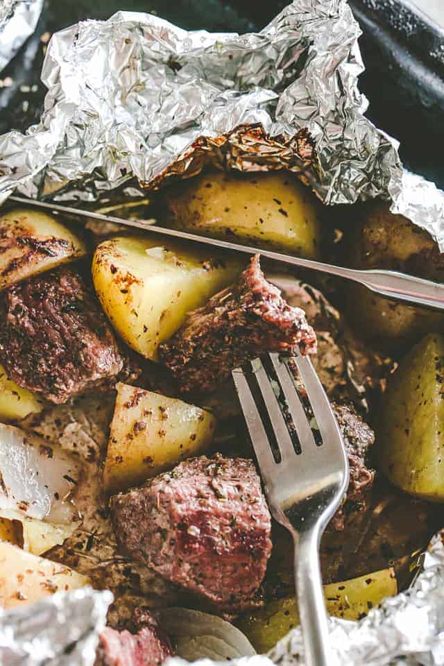 Garlic and herb steak and potatoes inside an open foil packet with a fork and knife.
