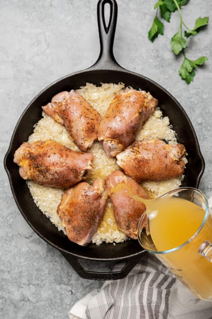 Pouring chicken broth over raw chicken thighs and rice in a skillet.