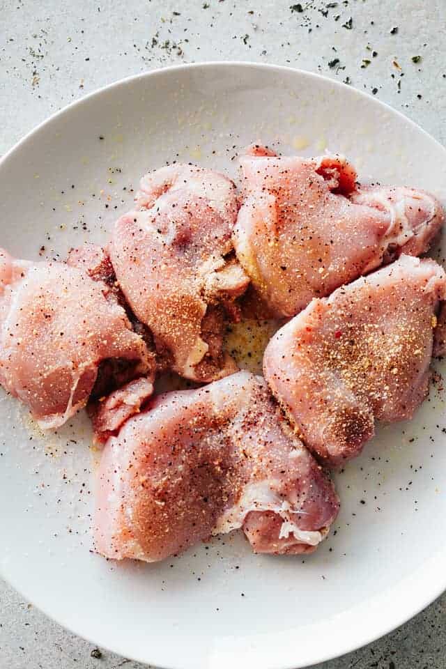 Seasoned raw boneless, skinless chicken thighs placed on a white plate.