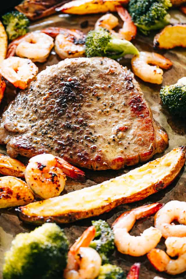 A close-up shot of shrimp, broccoli, a steak, and a potato wedge on an oiled baking sheet.