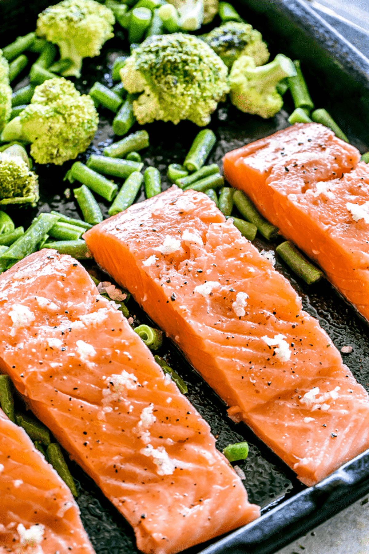 four raw salmon fillets arranged on a baking sheet with broccoli and green beans set above the fillets.