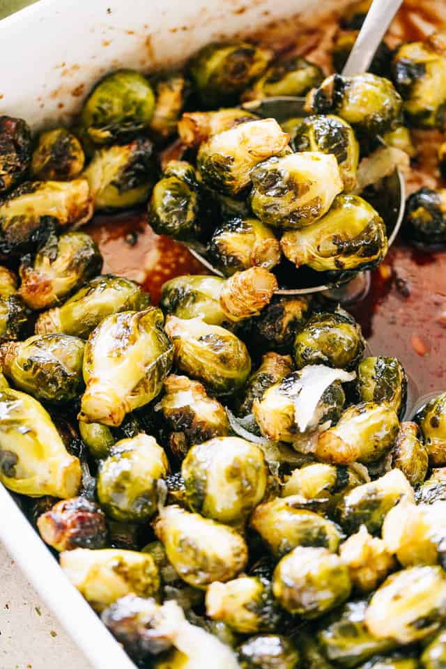Oven roasted Brussels sprouts in a white ceramic roasting pan next to a large spoon for serving.