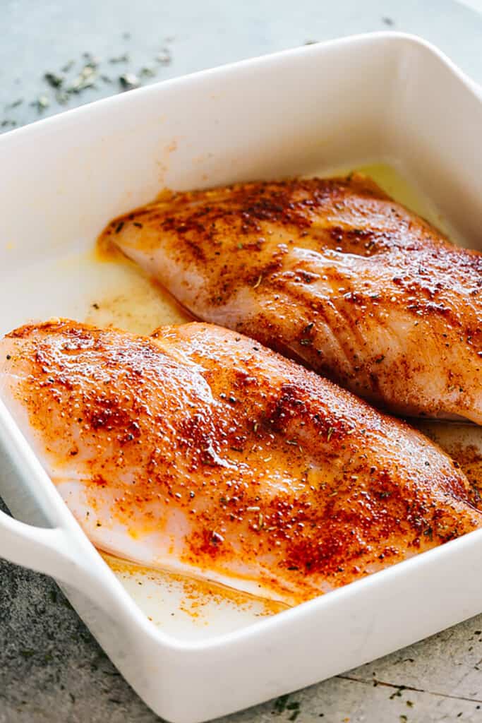 two raw chicken breasts, seasoned, and placed inside a white square baking dish.
