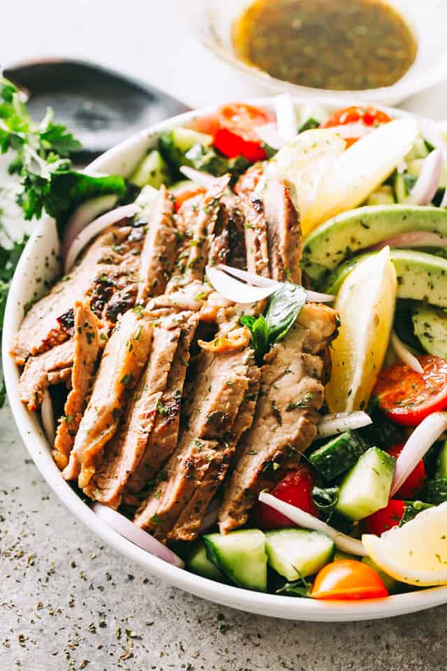 Pouring dijon balsamic dressing over a Steak Salad served in a large bowl with fresh veggies and lemon slices.