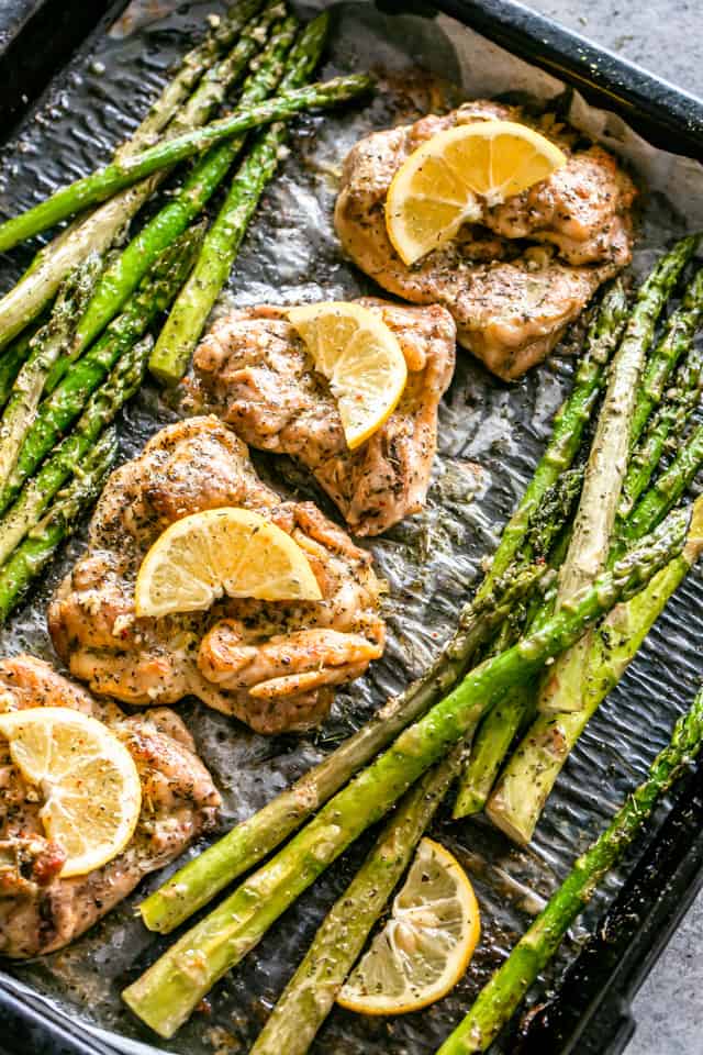 Overhead view of lemon garlic chicken thighs garnished with lemon slices, next to asparagus on a baking pan.
