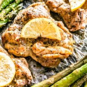 Close up overhead view of lemon garlic chicken thighs garnished with lemon slices, next to asparagus on a baking pan.