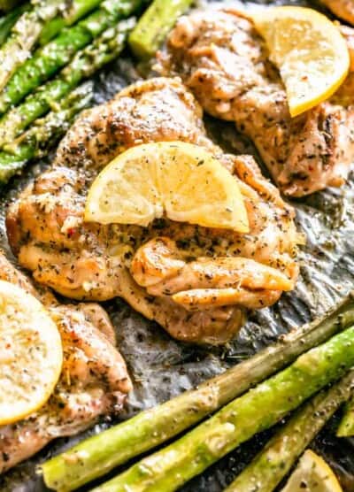 Close up overhead view of lemon garlic chicken thighs garnished with lemon slices, next to asparagus on a baking pan.