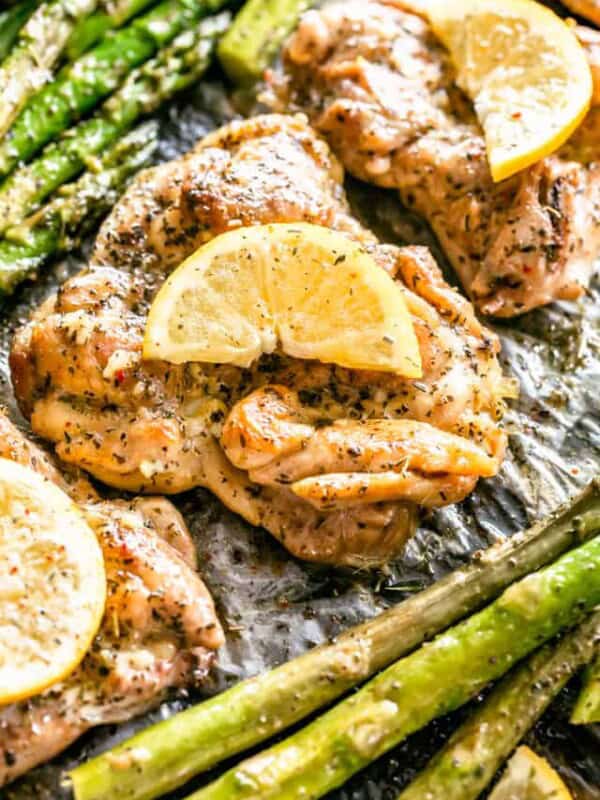 Close up overhead view of lemon garlic chicken thighs garnished with lemon slices, next to asparagus on a baking pan.